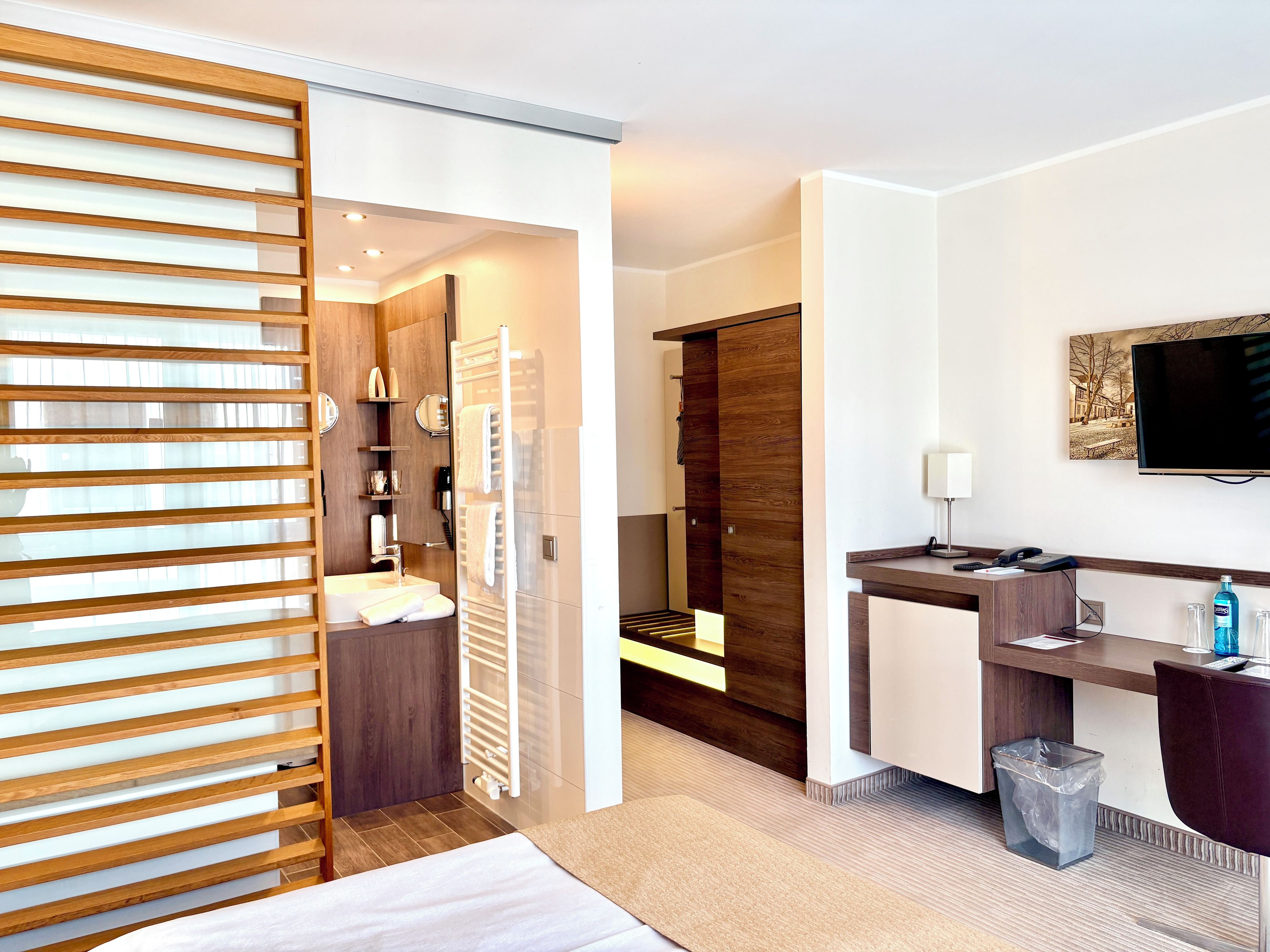 A modern hotel room with a wooden sliding door, a bathroom with a sink and mirror, a bedside table with a lamp, and a TV mounted on the wall.