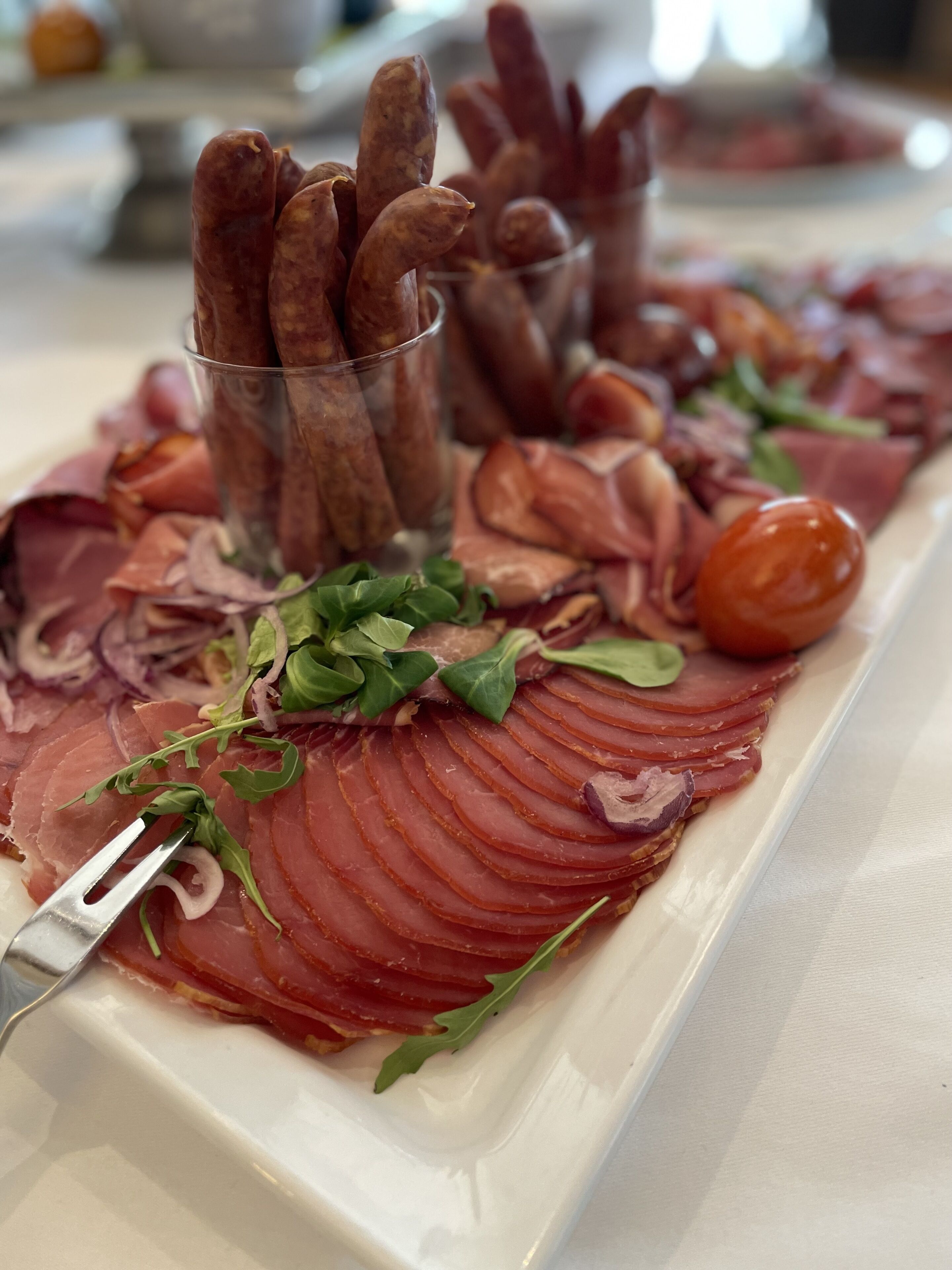 A platter of sliced meats and vegetables, including prosciutto, arugula, and cherry tomatoes, on a white plate.