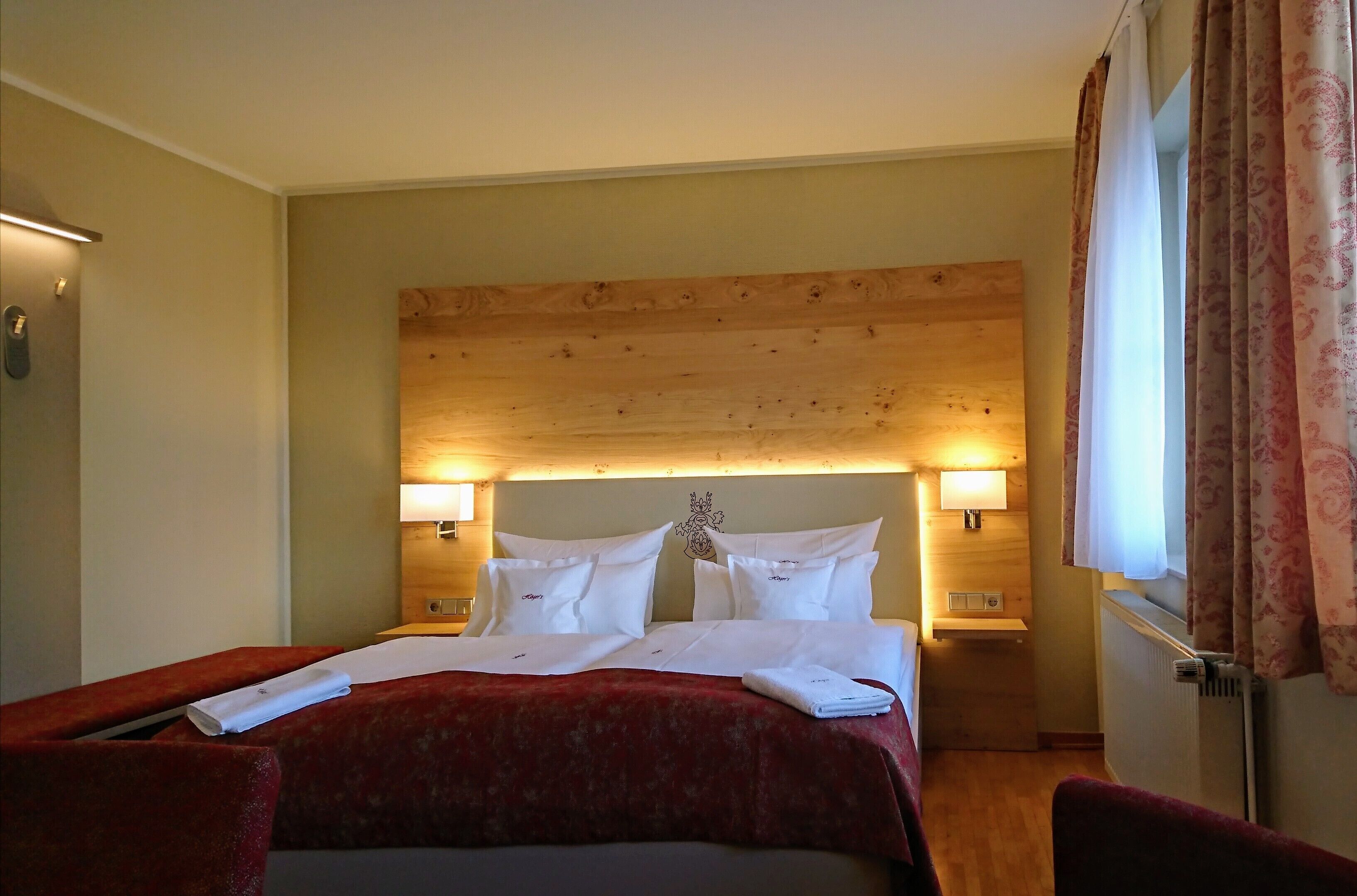 A hotel room with a wooden headboard, two bedside lamps, a red bedspread, and a window with curtains.