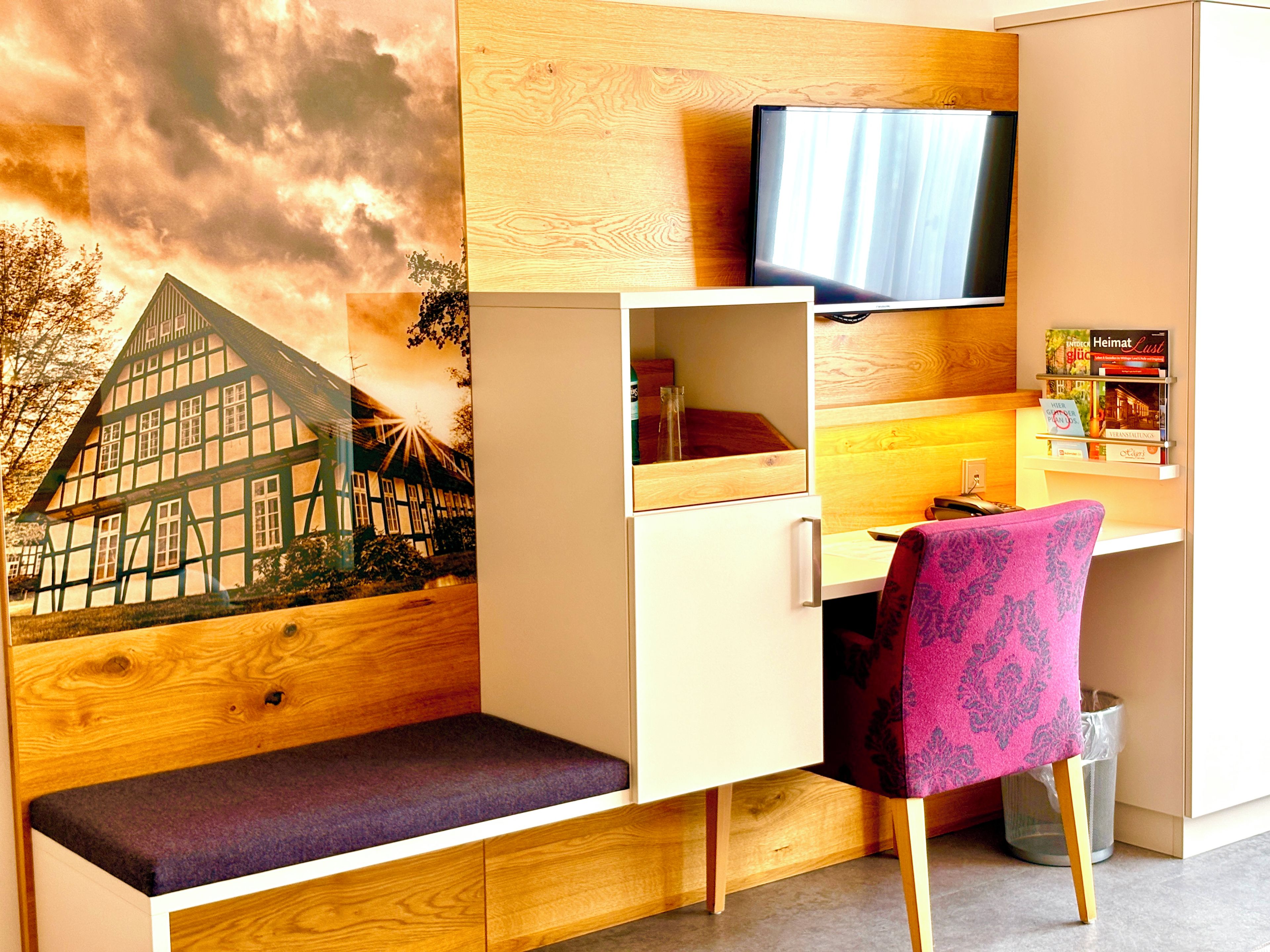 Una habitación con escritorio, televisión montada en la pared, estanter ía con libros y una banca.