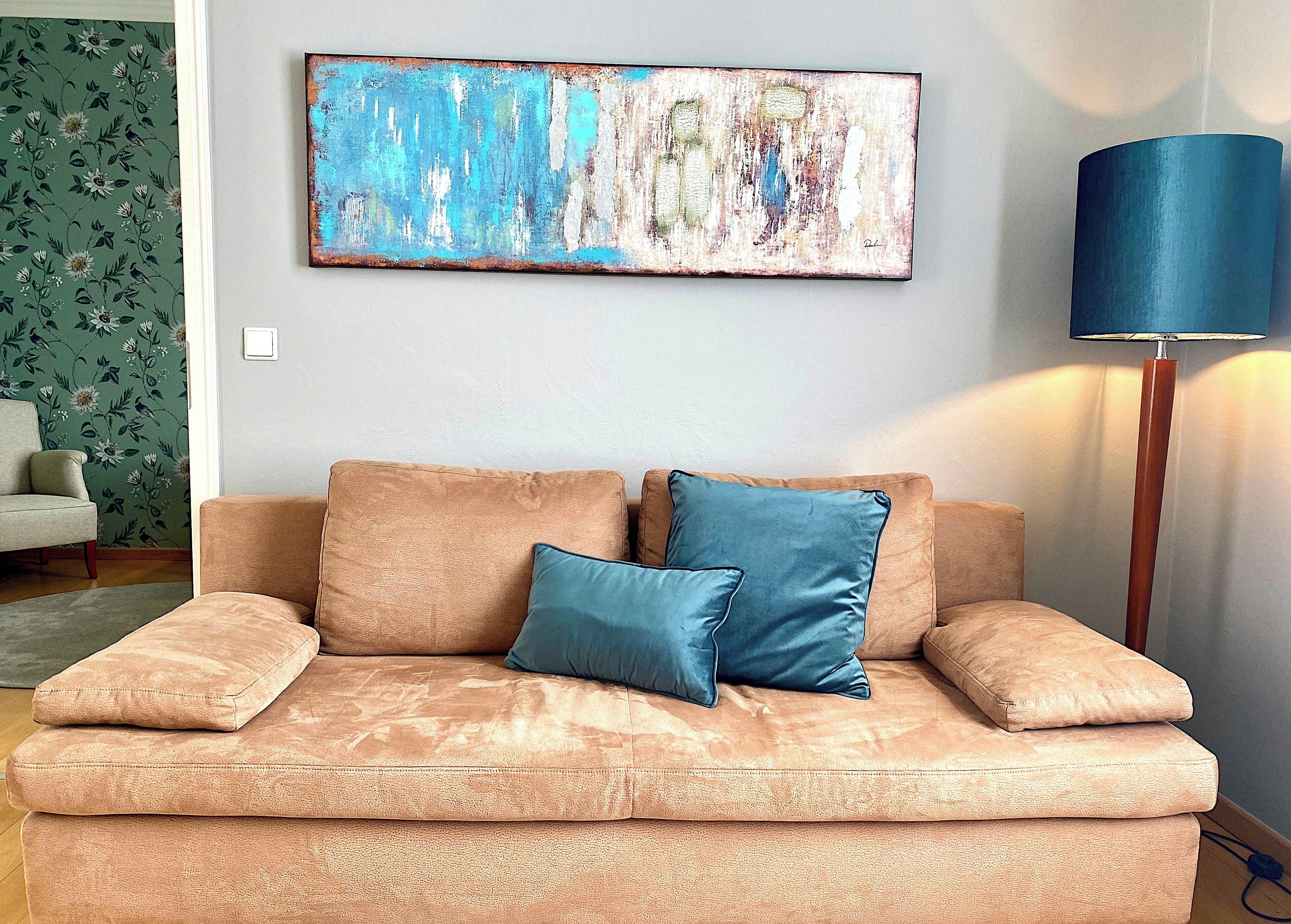 Un sofá color beige con dos cojines azules, una lámpara de pie azul y una pared cubierta de papel tapiz floral.