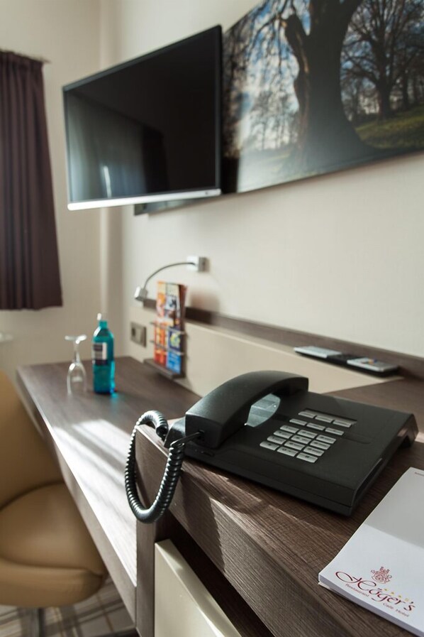 A hotel desk with a telephone, a glass of water, a bottle of hand sanitizer, a magazine rack, and a framed picture on the wall. - Högers Hotel und Restaurant (Bad Essen)