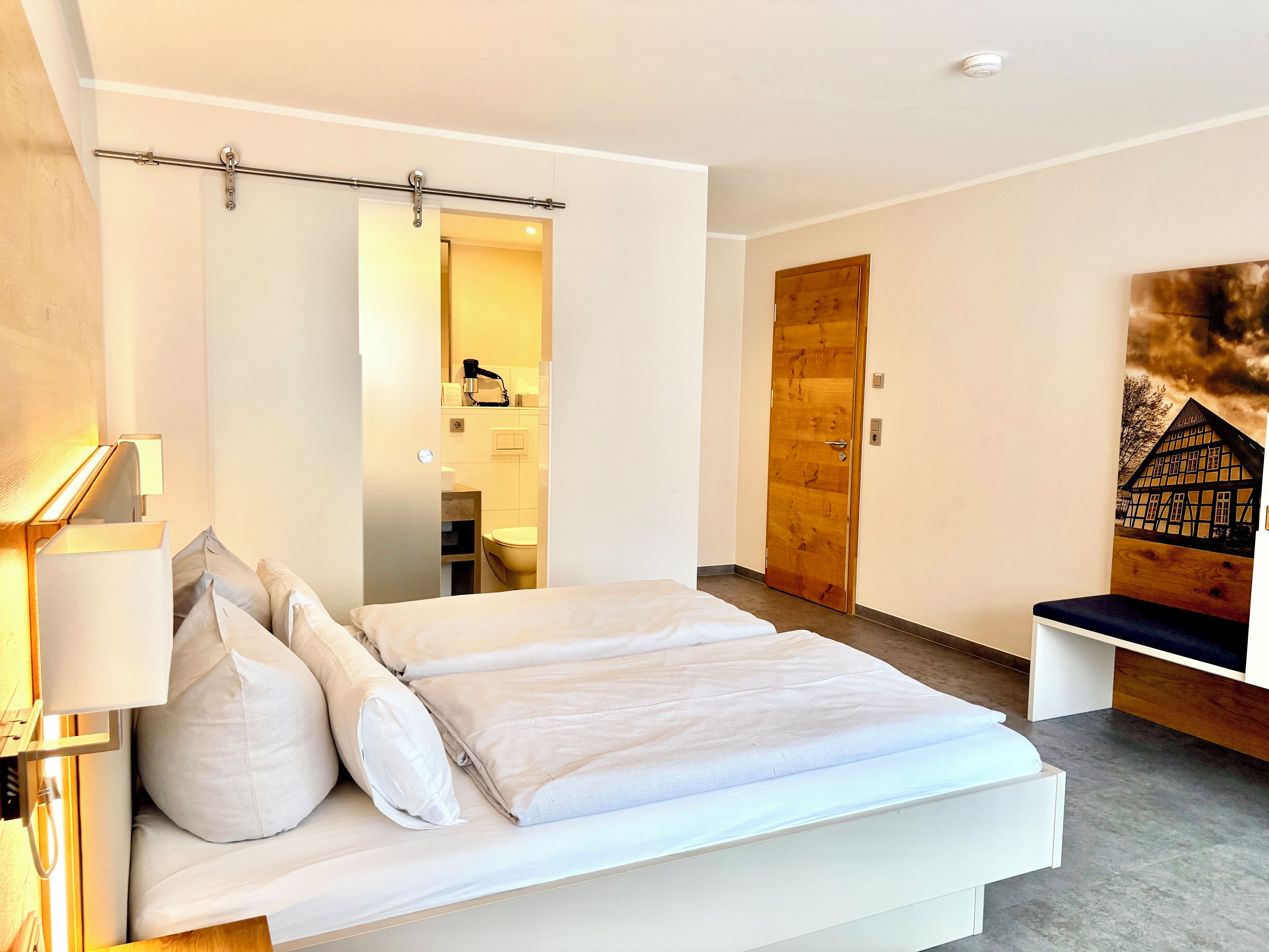 A modern bedroom with a large bed, a bedside lamp, a wooden door, and a wall-mounted artwork.