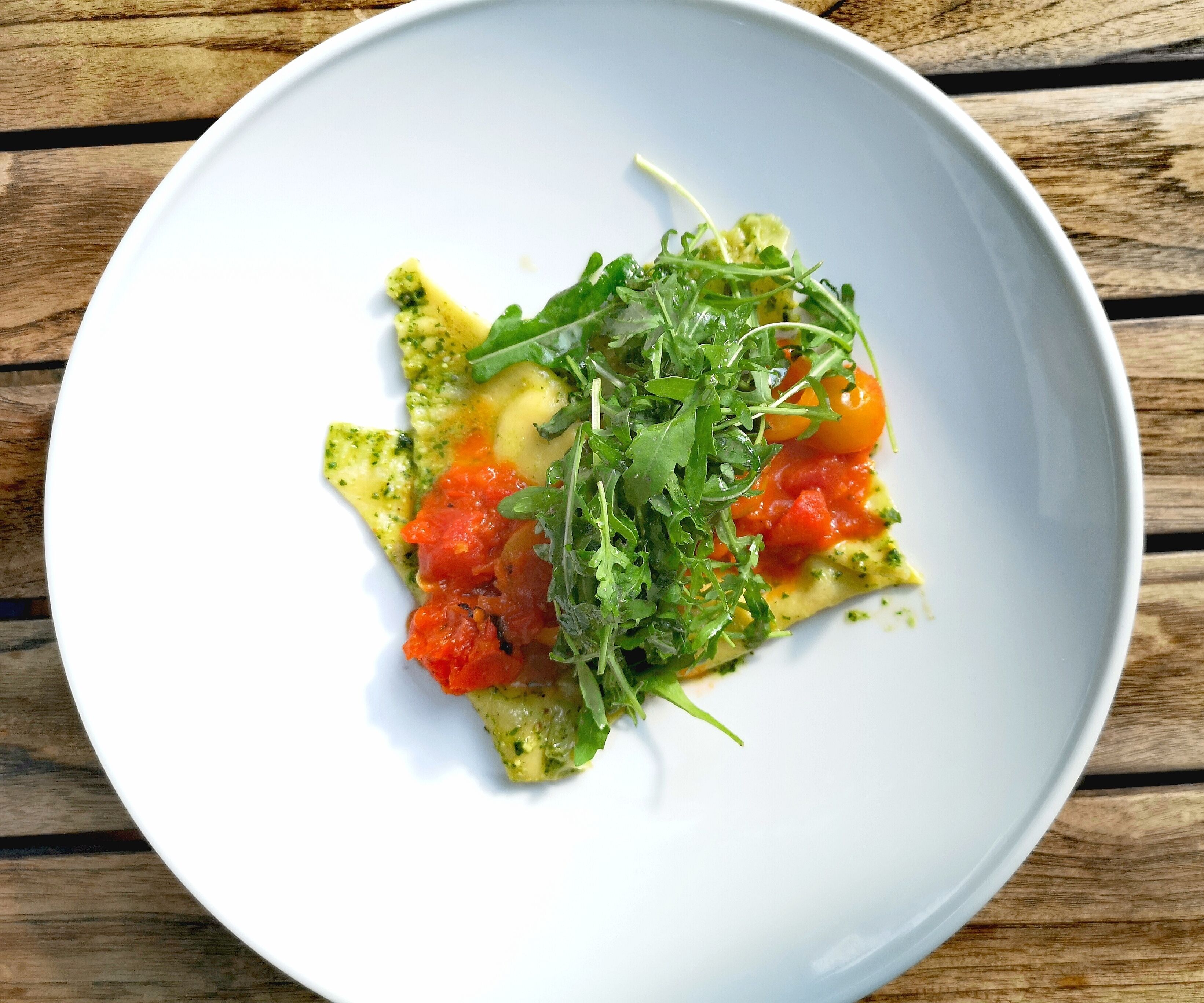 Un plato blanco con una porción de pasta, salsa de tomate y vegetales verdes sobre una superficie de madera.