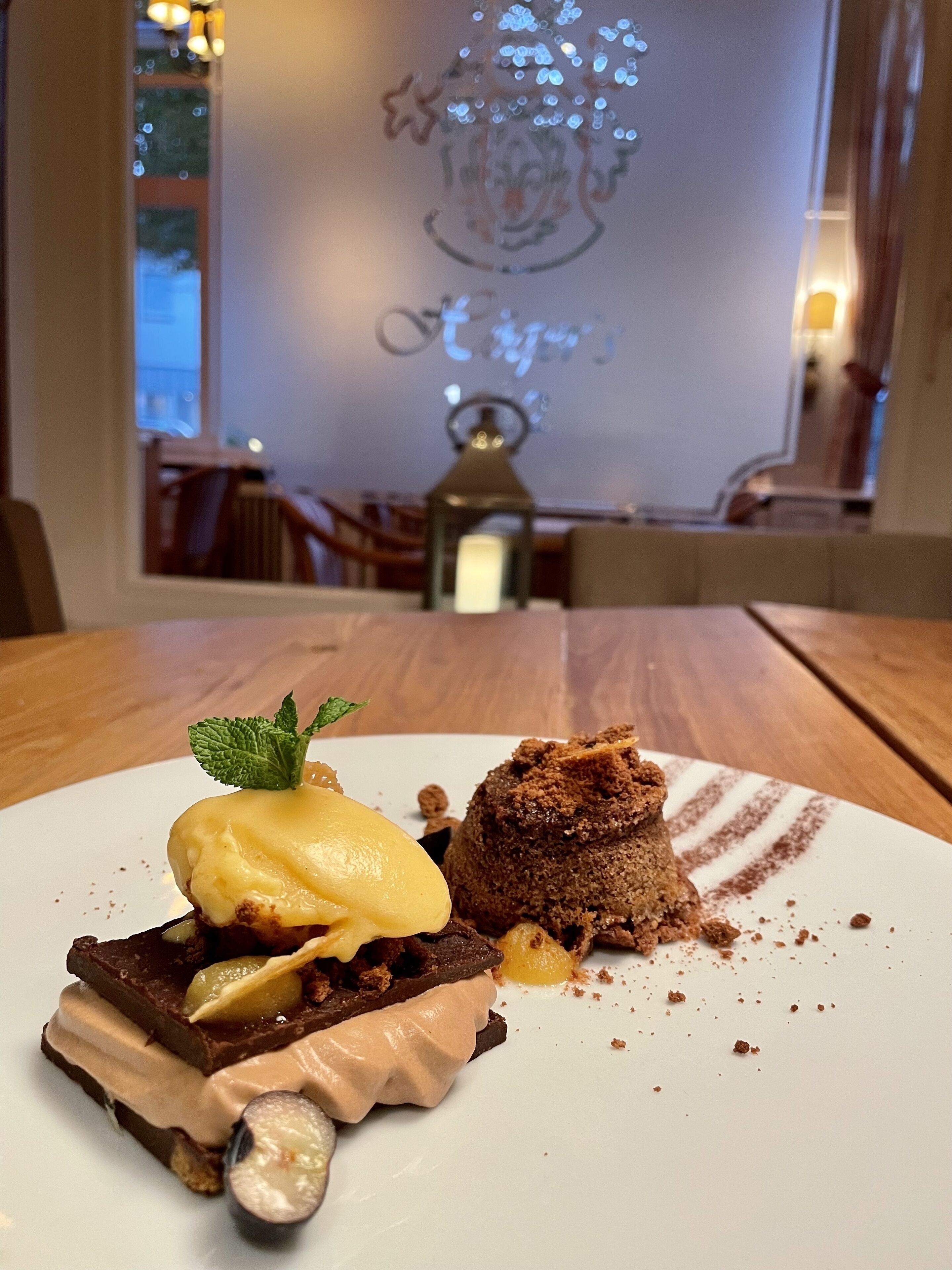 A dessert on a white plate with a mint leaf garnish, a chocolate cake, and a scoop of ice cream.