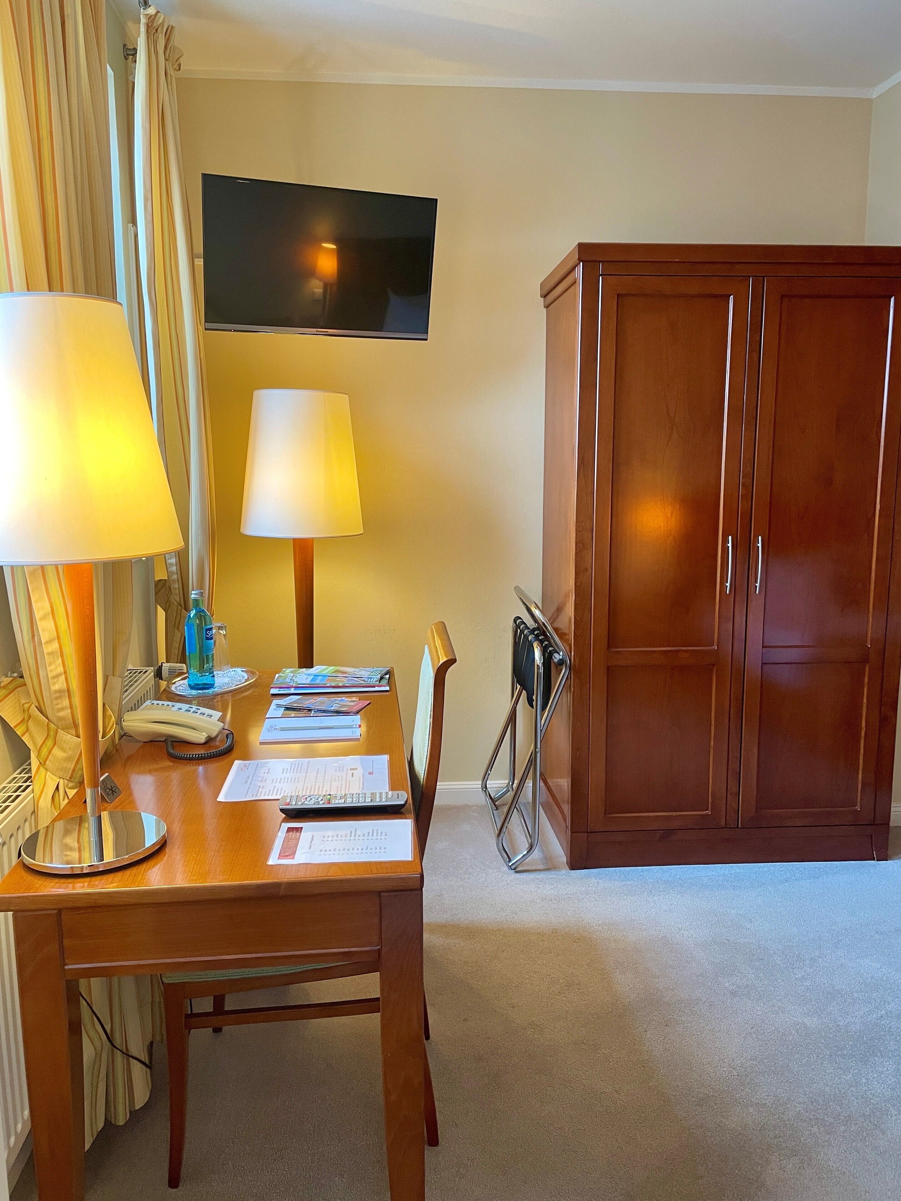 Habitación de hotel con escritorio de madera, dos lámparas, televisor, armario y radiador.