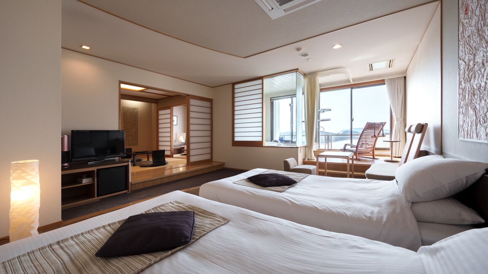 A hotel room with two beds, a TV, a desk, and a view of the outside.