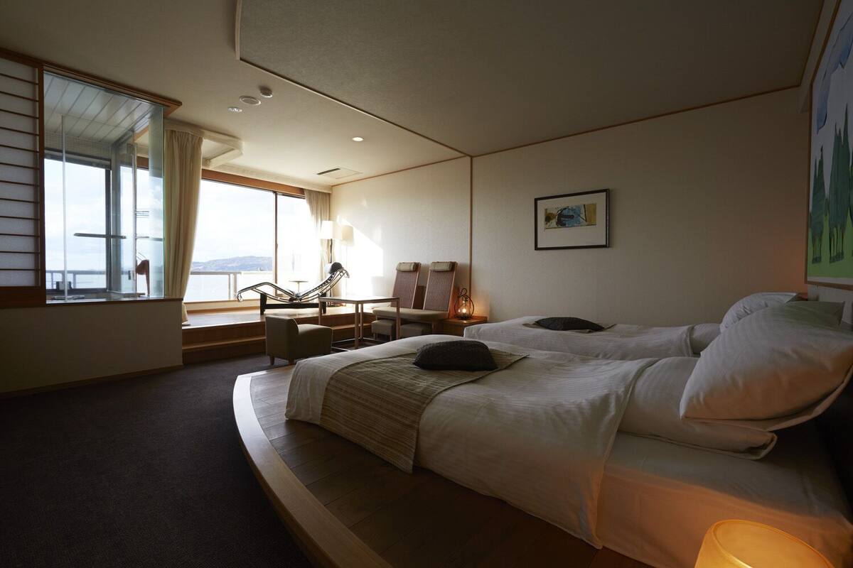 A hotel room with two beds, a sitting area with a chair and table, a large window with curtains, and a painting on the wall.