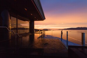 Eine Terrasse am Wasser mit Glasgeländer, einer Metalltreppe und Blick auf den Sonnenuntergang über dem Wasser.