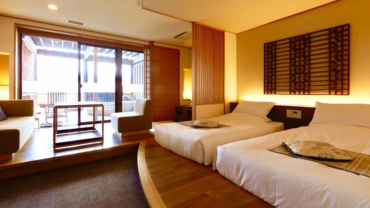 A hotel room with two beds, a sofa, a table, and a view of the outside.