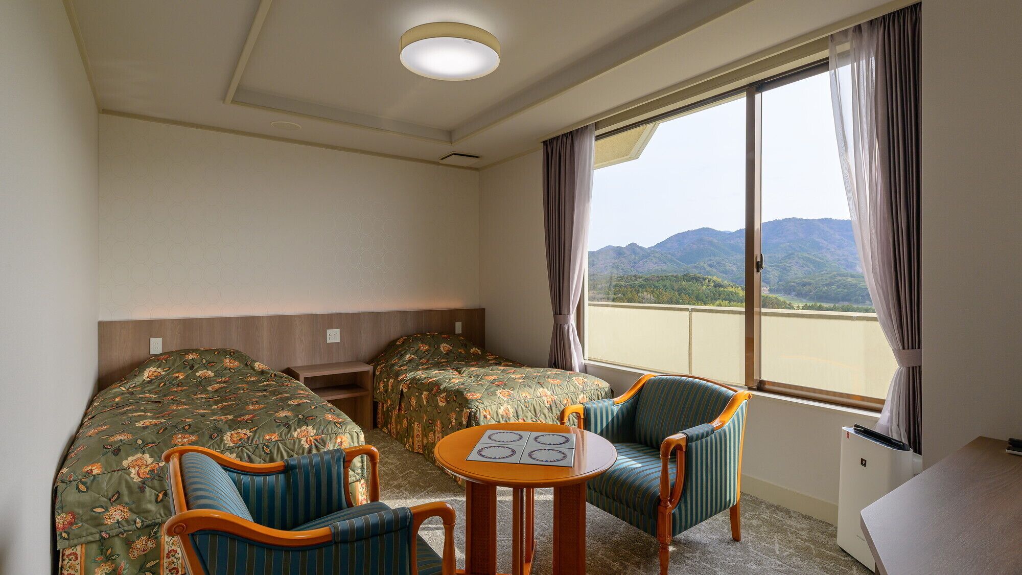 A hotel room with two beds, a small table, two chairs, a window with a view of mountains, and a ceiling light.