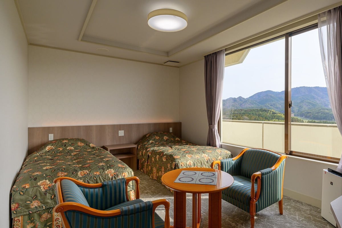 Une chambre d’hôtel avec deux lits, une petite table, deux chaises, une fenêtre donnant sur les montagnes et un luminaire au plafond.
