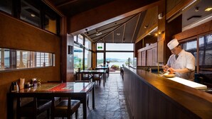 A restaurant with a long counter, wooden tables, and chairs. A chef is preparing food at the counter. - Yuyawan Onsen Hotel Yokikan (Nagato)