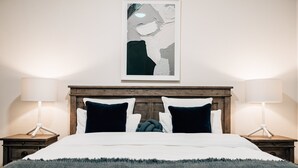 Ein ordentlich bezogenes Bett mit einem hölzernen Kopfteil, zwei Nachttische mit Lampen und ein gerahmtes abstraktes Kunstwerk darüber.