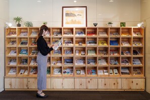 A woman browsing books in a wooden bookcase with multiple shelves, in a room with a tiled wall and a framed picture above the shelves.