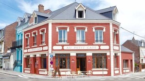 A red building with the sign "AU PETIT CAFE" on the front. There are outdoor tables and chairs in front of the cafe. - Hidden Bay Hotel (Cayeux-sur-Mer)