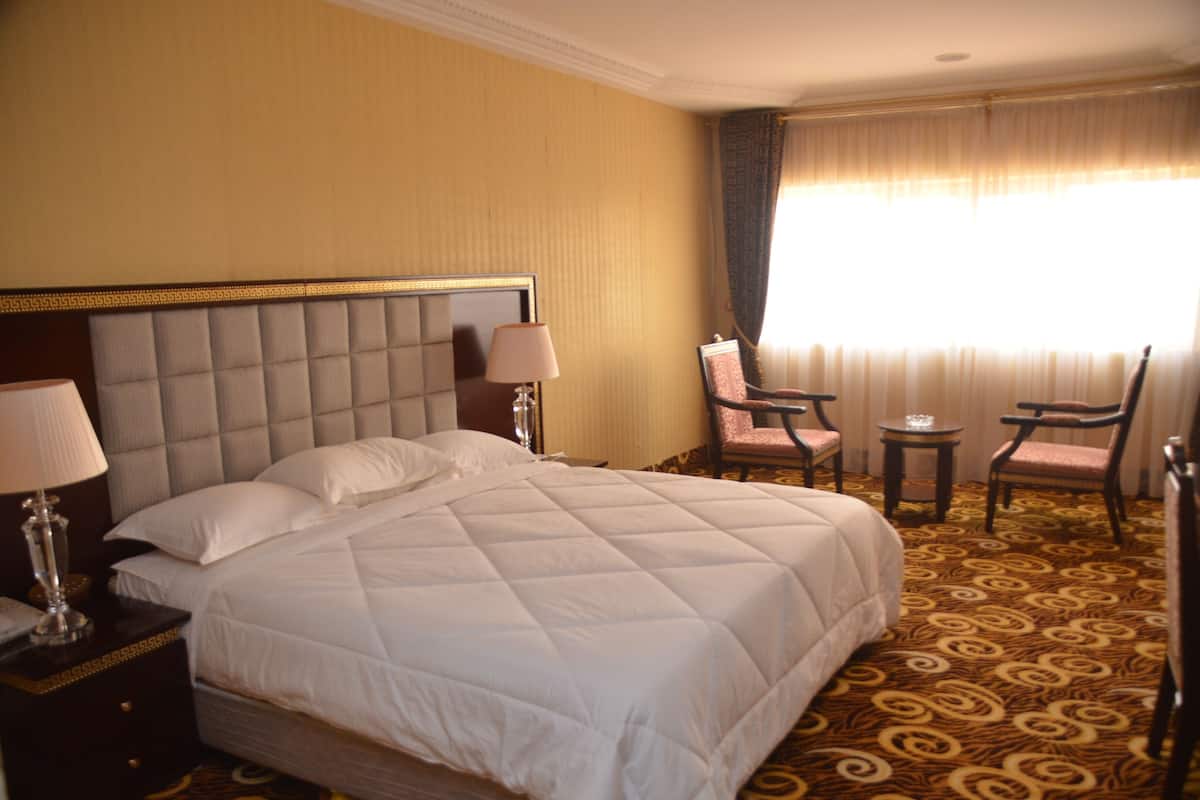 Ein Hotelzimmer mit einem großen Bett, zwei Nachttischen mit Lampen, einem kleinen Tisch mit einem Stuhl und einem großen Fenster mit Vorhängen.