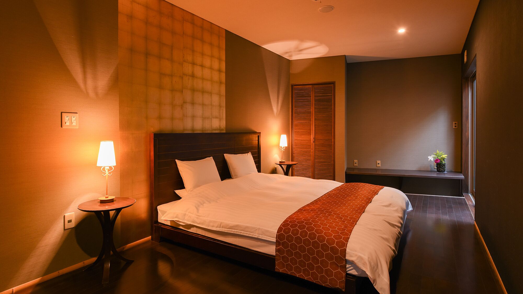 Een hotelkamer met een groot bed, nachtlampjes, een houten hoofdbord, een klein tafeltje en een nachtkastje.