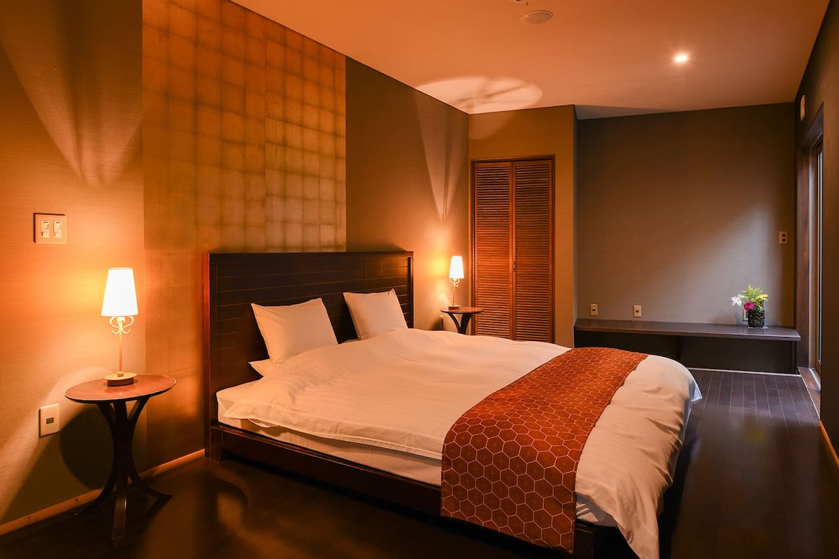 Habitación de hotel con una cama grande, lámparas de noche, cabecero de madera, una mesita pequeña y un buró.