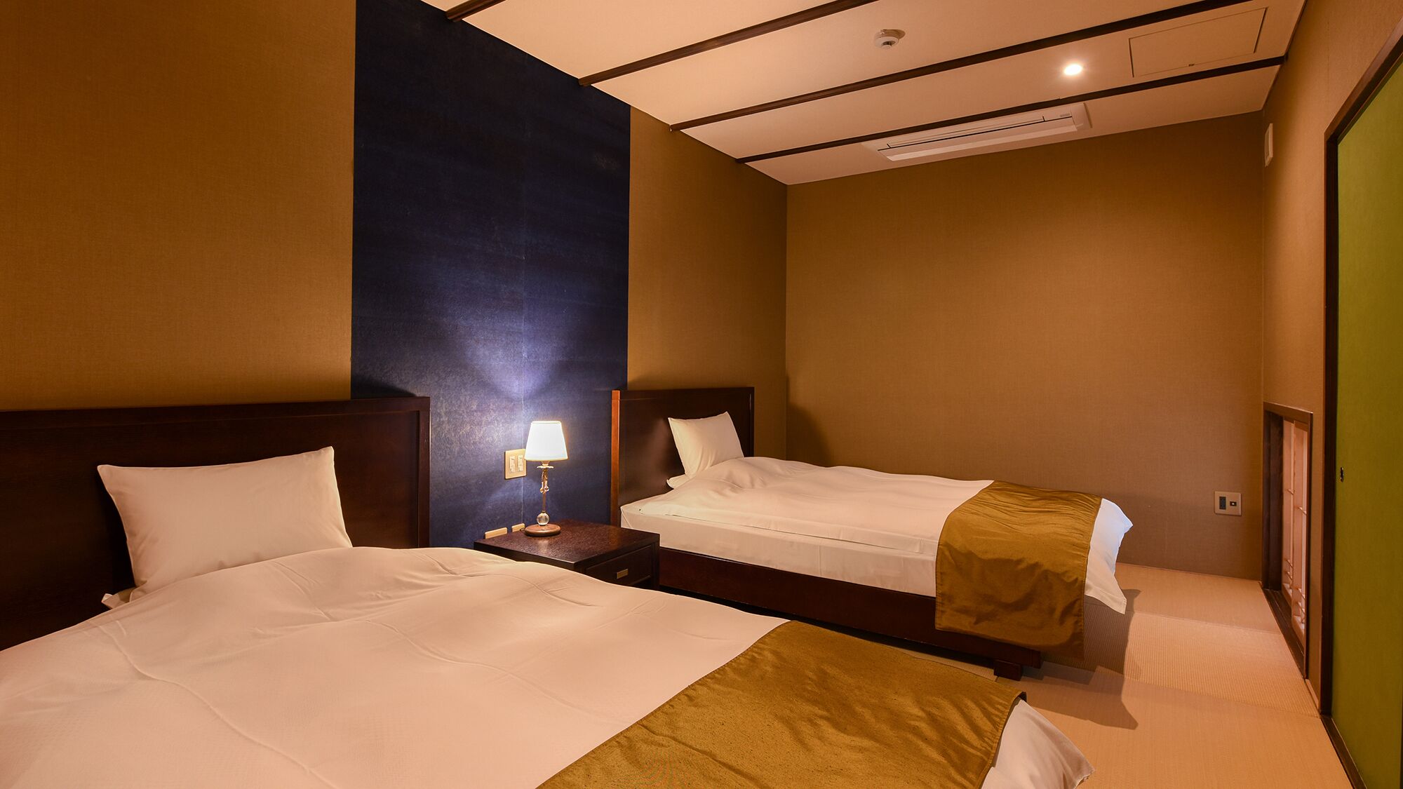 Een hotelkamer met twee bedden, een nachtkastje met een lamp en een plafond met inbouwverlichting.