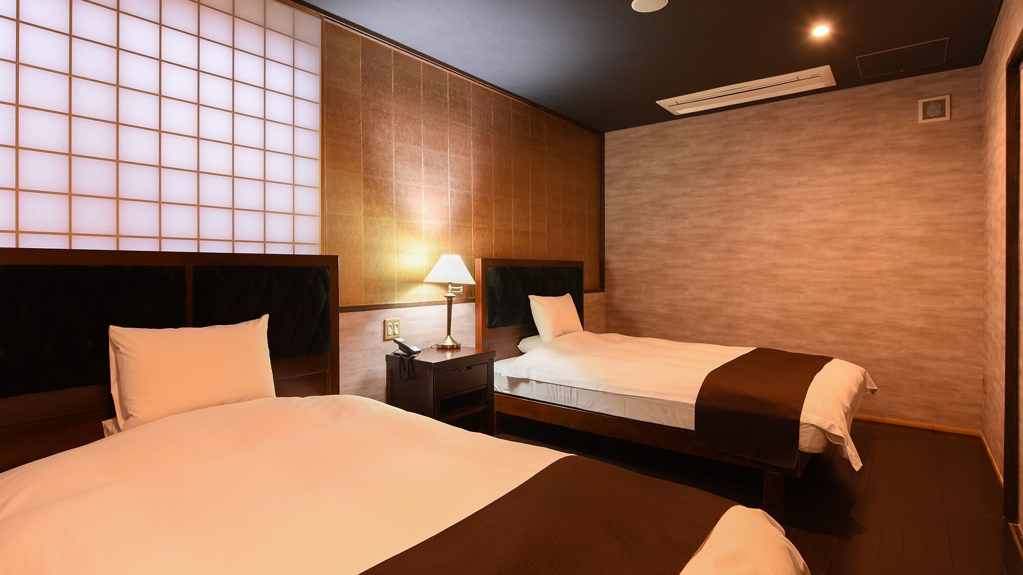 Een hotelkamer met twee bedden, een nachtkastje met een lamp en een groot raam met een rasterpatroon.