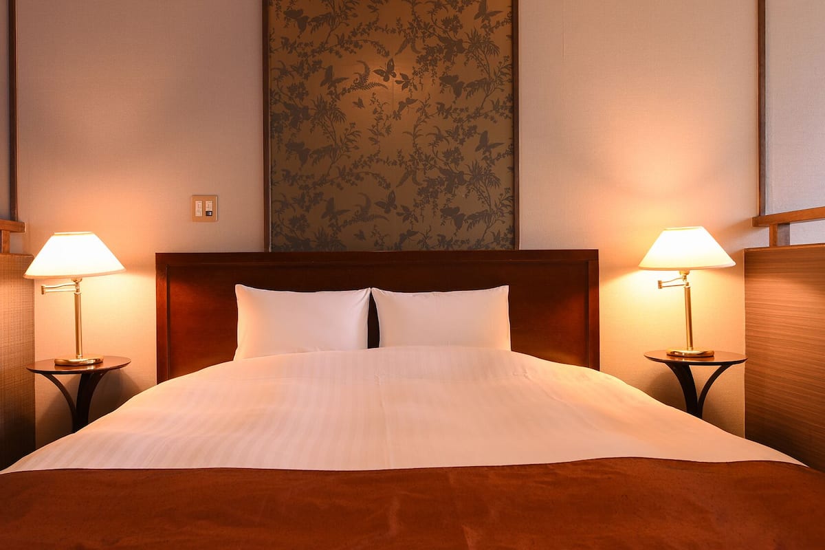 Habitación de hotel con una cama grande, dos lámparas de noche, cabecera de madera y papel tapiz con motivos florales.