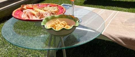 Una mesa de vidrio con un plato de comida y un vaso de bebida roja, ubicada en un balcón con vistas a una ladera y edificios.