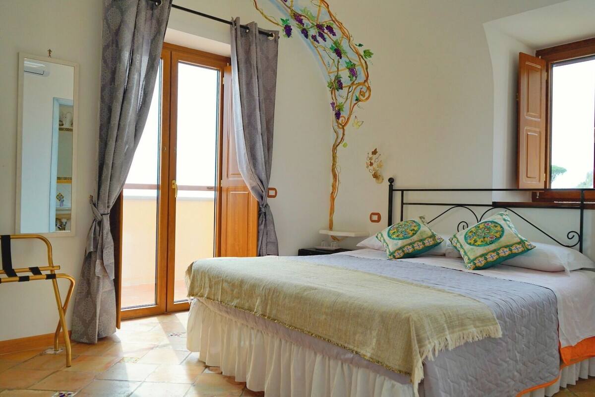 A bedroom with a bed, curtains, a nightstand, a wall-mounted shelf, and a window with a view.