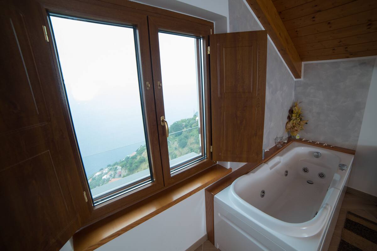 A bathroom with a bathtub, wooden cabinets, and a large window offering a view of the sea.