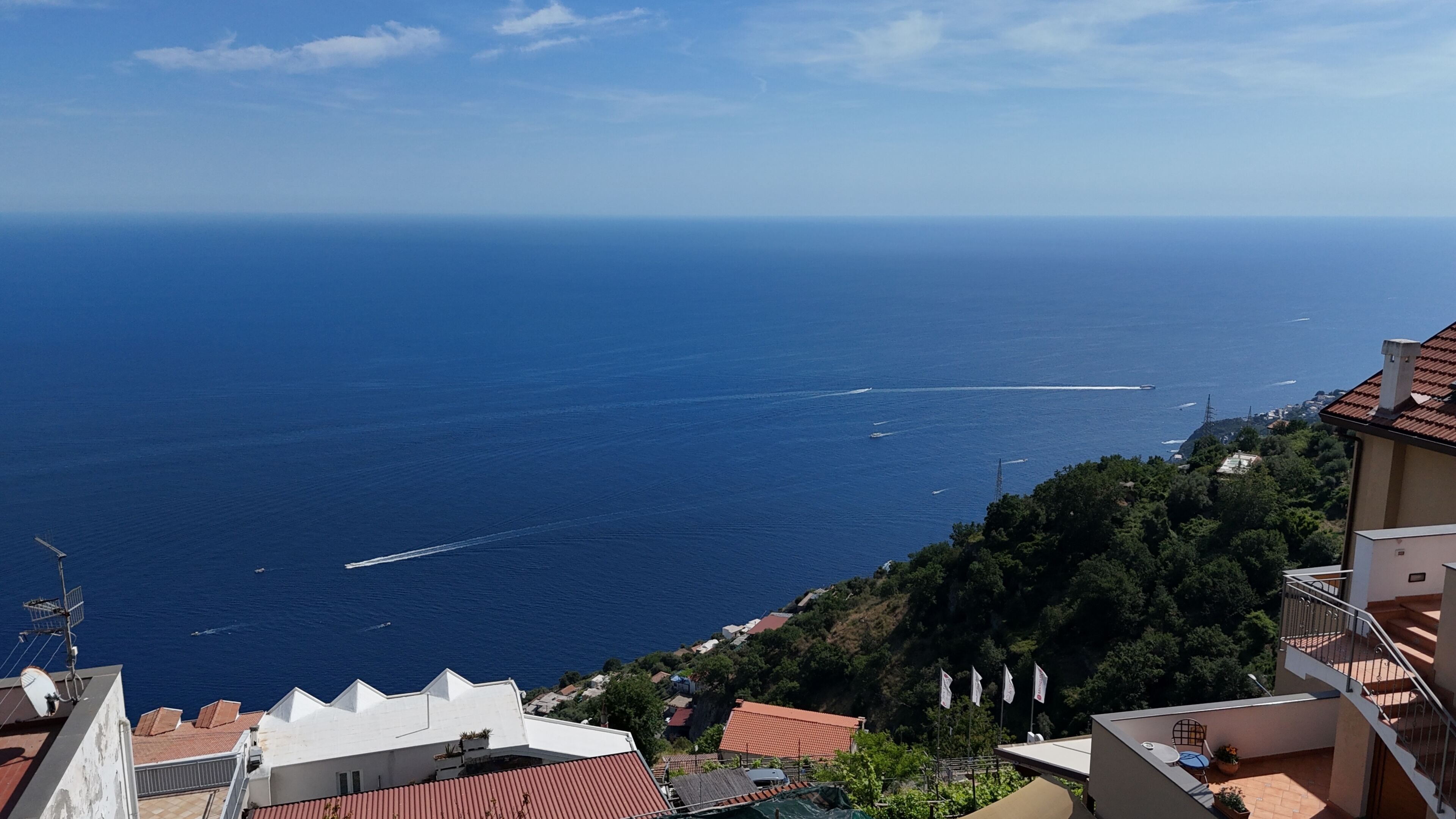 A coastal view with a clear sky, a boat creating a wake, and buildings with red-tiled roofs.