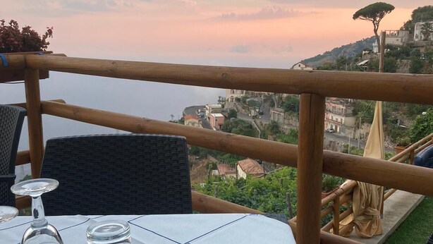 A table set with cutlery and glasses, overlooking a coastal town with buildings and a tree on a hill. - San Giacomo Relais (Furore)