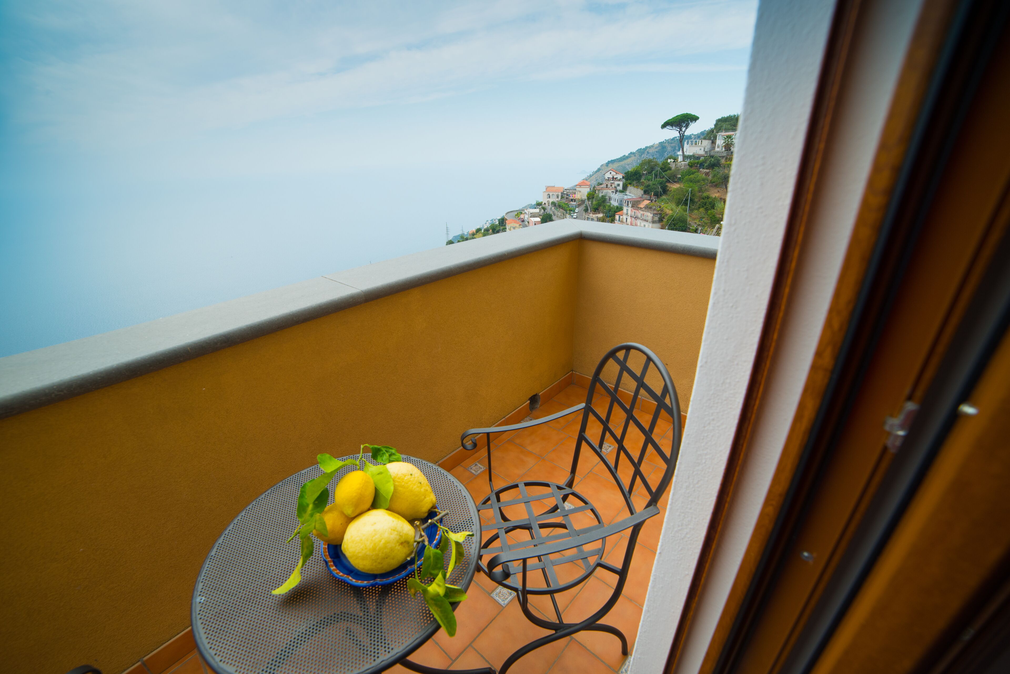 A balcony with a table and chair, a bowl of lemons, and a view of a hillside with buildings.