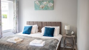 Una cama bien hecha con almohadas azules y blancas, una mesita de noche con una lámpara y un despertador, y una obra de arte floral en la pared.