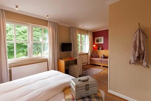 A hotel room with a bed, a desk with a television, a chair, a sofa, and a window with curtains.