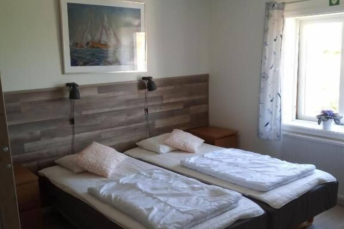 Ein Zimmer mit zwei Betten, einem Bild an der Wand, einer Deckenleuchte und einem Fenster mit Vorhängen.