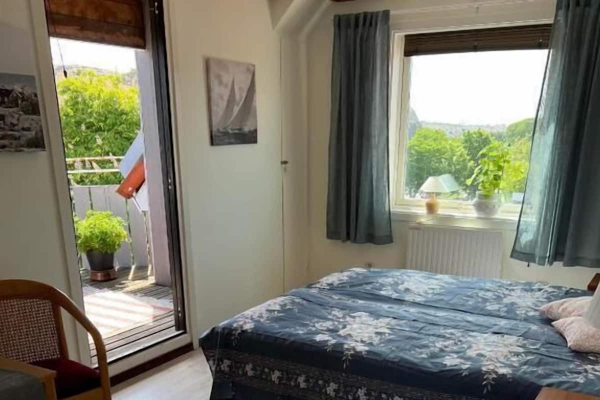 Ein Schlafzimmer mit Bett, Stuhl, einem Fenster mit Vorhängen und Blick auf Grün.
