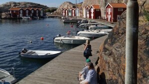 Eine hölzerne Pier mit angestaubten Booten, eine felsige Küstenlinie und rote Gebäude mit weißen Verzierungen.