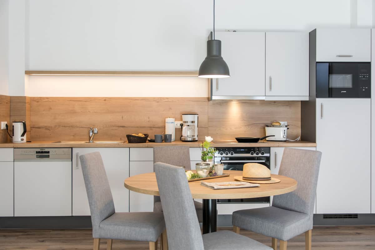 Cocina moderna con armarios blancos, encimera de madera, mesa redonda para comer y sillas tapizadas en gris.