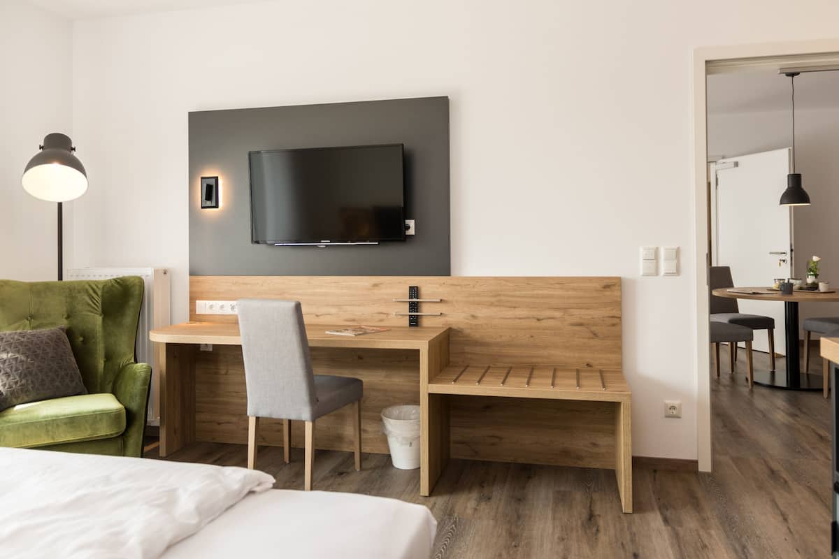 Una habitación de hotel moderna con un escritorio de madera, un televisor de pantalla plana montado en la pared, un sillón verde y una cama con ropa de cama blanca.