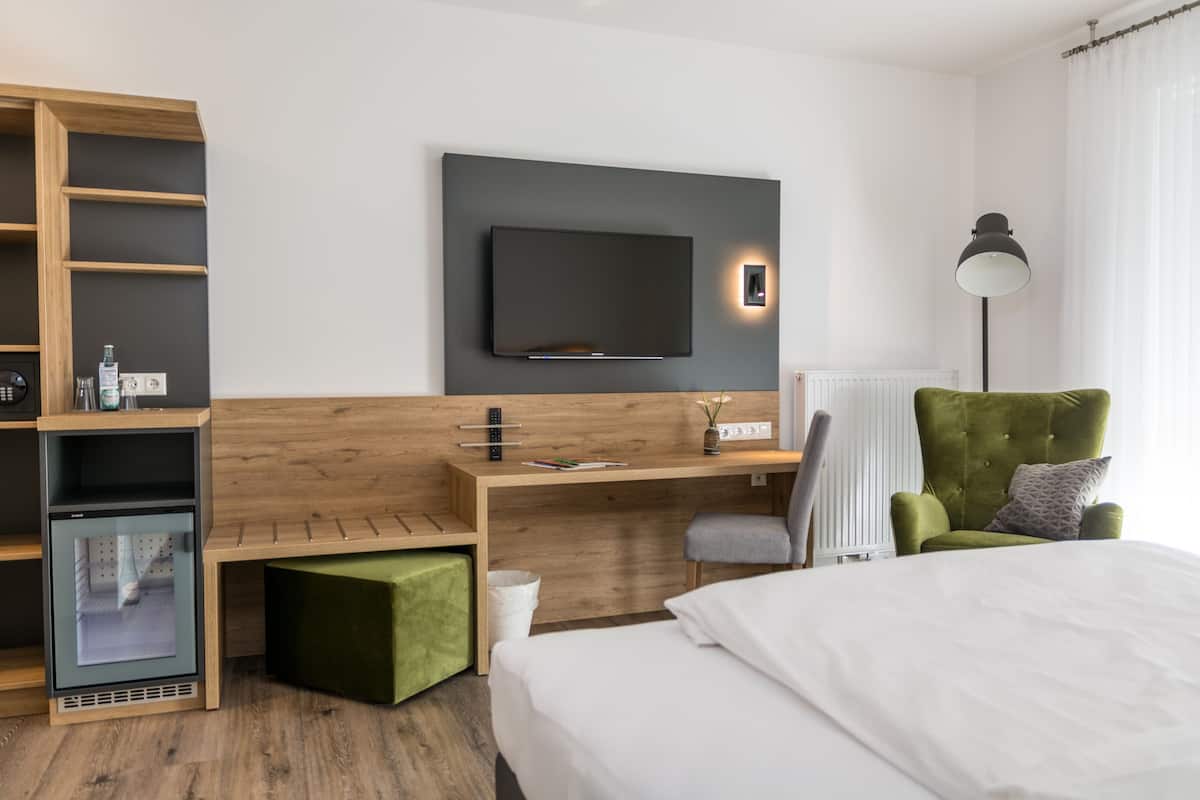 Una habitación de hotel moderna con cama, escritorio con televisor de pantalla plana, sillón verde y un mini refrigerador integrado.