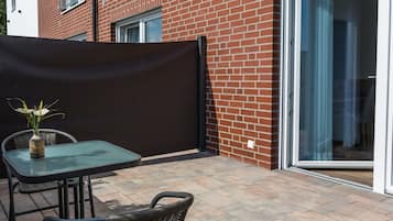 A patio with a glass table and chairs, a wicker chair, a potted plant, and a brick building with a sliding glass door.