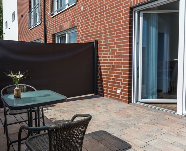 Une terrasse avec une table et des chaises en verre, un fauteuil en osier, une plante en pot et un bâtiment en briques doté d’une porte-fenêtre coulissante.