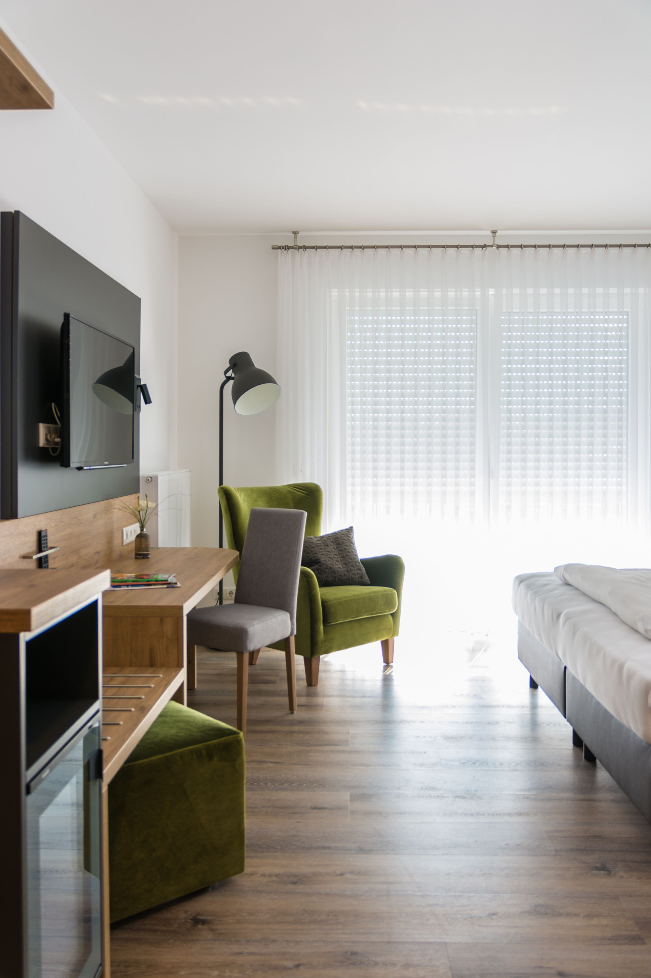 Une chambre d’hôtel moderne avec un bureau en bois, un fauteuil vert, un canapé gris, un téléviseur fixé au mur et une grande fenêtre avec des stores.