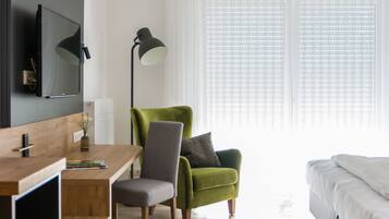 Une chambre d’hôtel moderne avec un bureau en bois, un fauteuil vert, un canapé gris, un téléviseur fixé au mur et une grande fenêtre avec des stores.