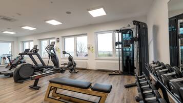 Academia com diversos equipamentos de exercício, amplas janelas e piso de madeira.