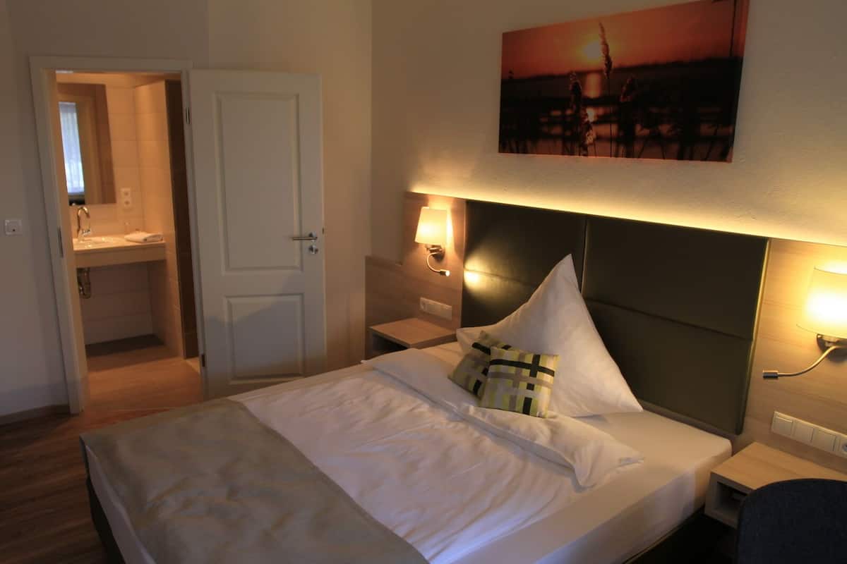 Una habitación de hotel con cama, mesitas de noche, una lámpara de pared y una puerta que da al baño.