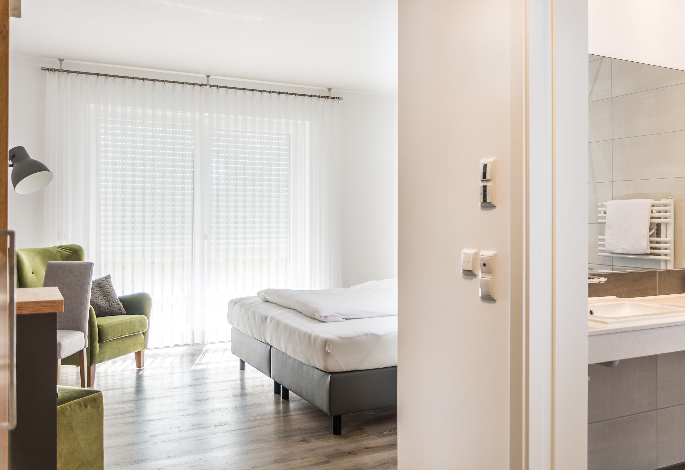 Een moderne hotelkamer met een bed, een groene fauteuil, een bureau en een badkamer met wastafel en spiegel.