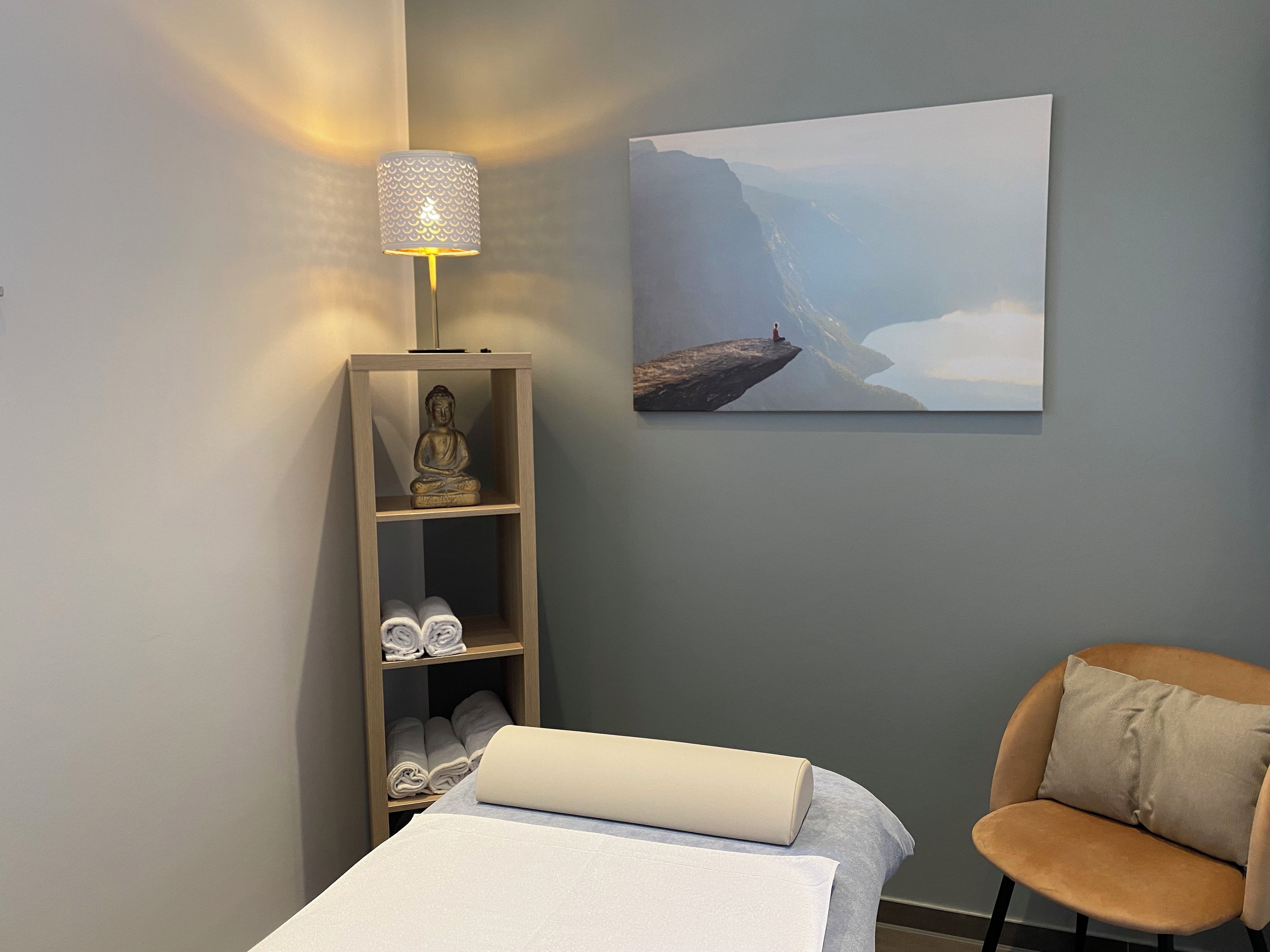 A room with a bed, a chair, a shelf with towels, a Buddha statue, a lamp, and a painting of a mountain landscape.