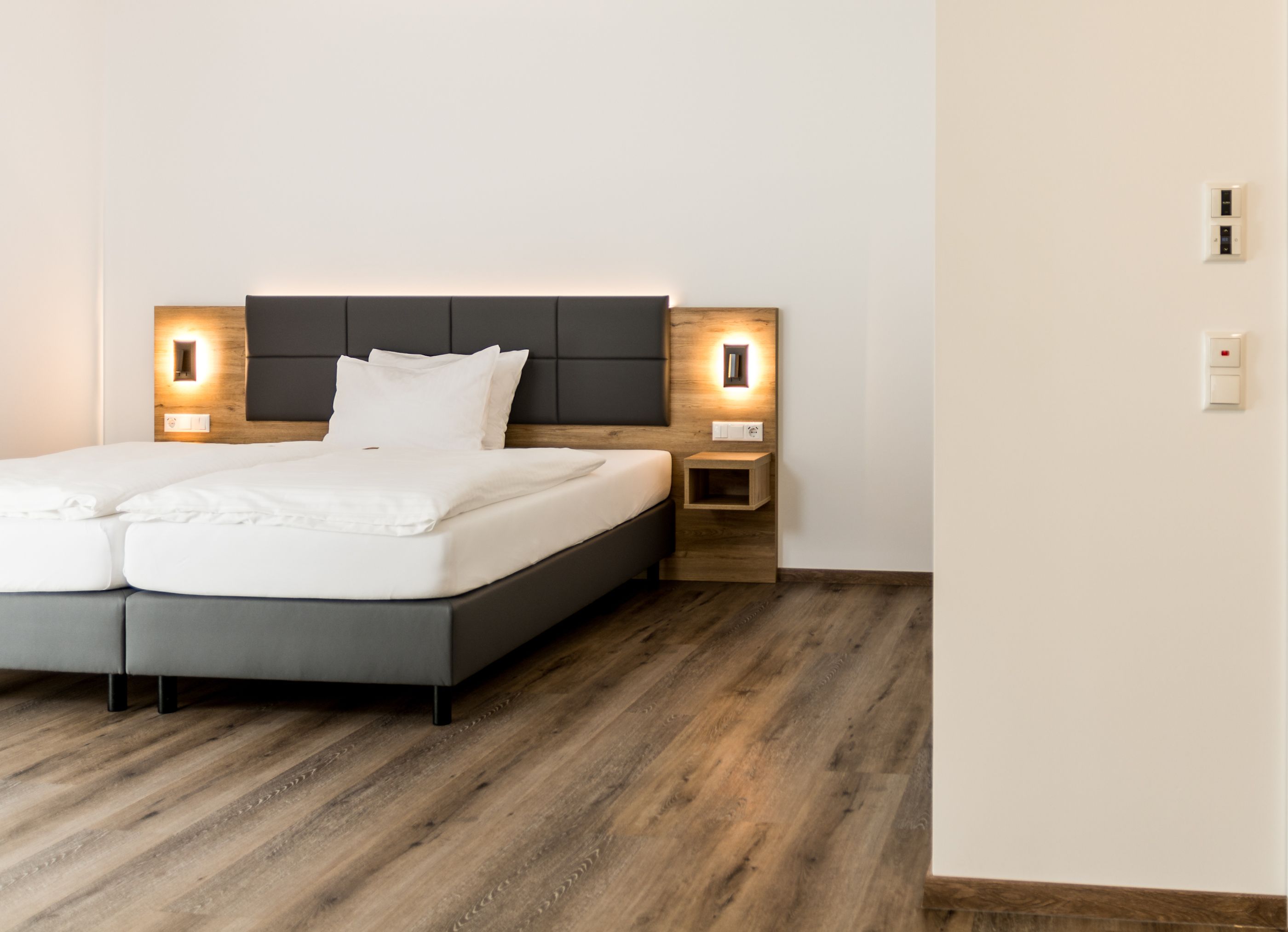 Ein modernes Schlafzimmer mit grauem Bett, Holz-Kopfteil, Nachttischen mit integrierter Beleuchtung und Holzboden.