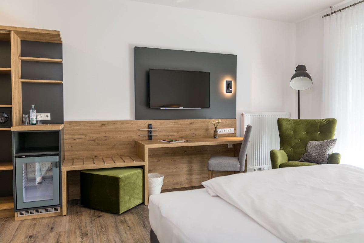 Une chambre d’hôtel moderne équipée d’un lit, d’un bureau avec un téléviseur à écran plat, d’un fauteuil vert et d’un mini-réfrigérateur intégré.